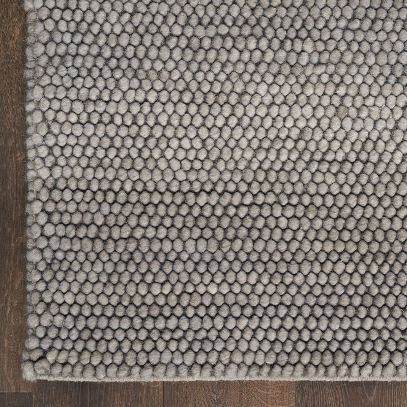 A close-up of a textured area rug with a dense, circular tufted pattern in a light gray color, shown on a dark brown wooden floor.
