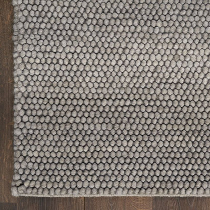 A close-up of a textured area rug with a dense, circular tufted pattern in a light gray color, shown on a dark brown wooden floor.