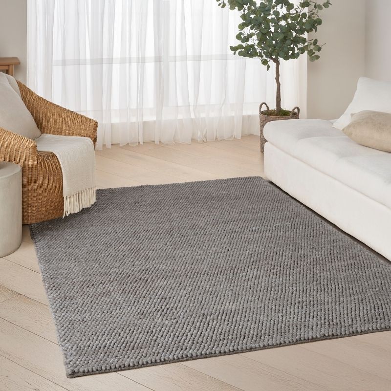 A large rectangular gray textured area rug laid on a light wood floor in a bright living room, with a white sofa, woven armchair, sheer curtains, and a potted tree visible.