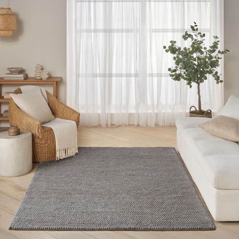 A gray textured woven area rug styled in a bright, modern living room with a white sofa, a wicker armchair, and sheer white curtains.