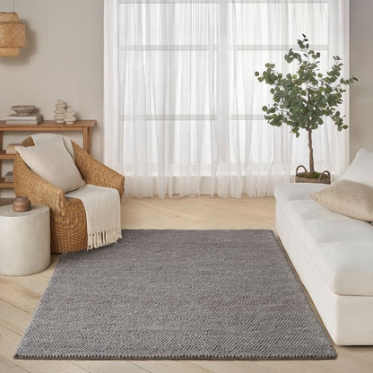 A gray textured woven area rug styled in a bright, modern living room with a white sofa, a wicker armchair, and sheer white curtains.
