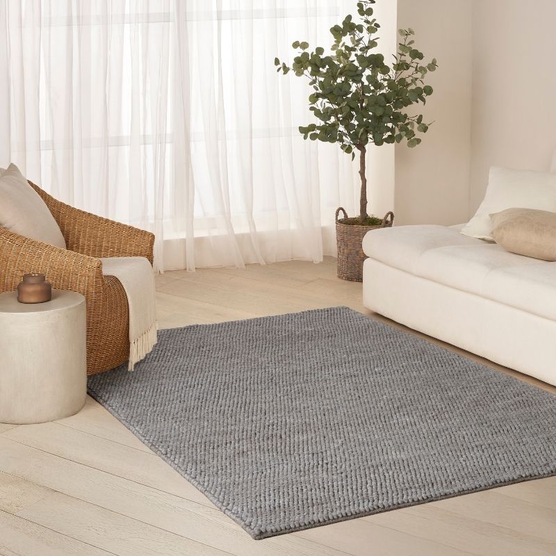 A rectangular gray woven area rug laid on a light wood floor in a bright, modern living room with a white sofa, wicker chair, side table, sheer curtains, and a potted tree.