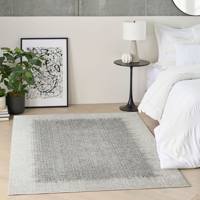 A rectangular area rug in a bedroom setting, featuring a distressed gray and white textured pattern. The rug is styled in a modern bedroom next to a bed with white bedding, a black side table with a lamp, a potted plant, and framed wall art.