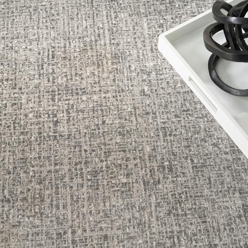 A close-up of a textured gray and beige woven fabric or carpet surface, with a white tray and black decorative object partially visible in the upper right corner.