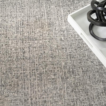 A close-up of a textured gray and beige woven fabric or carpet surface, with a white tray and black decorative object partially visible in the upper right corner.