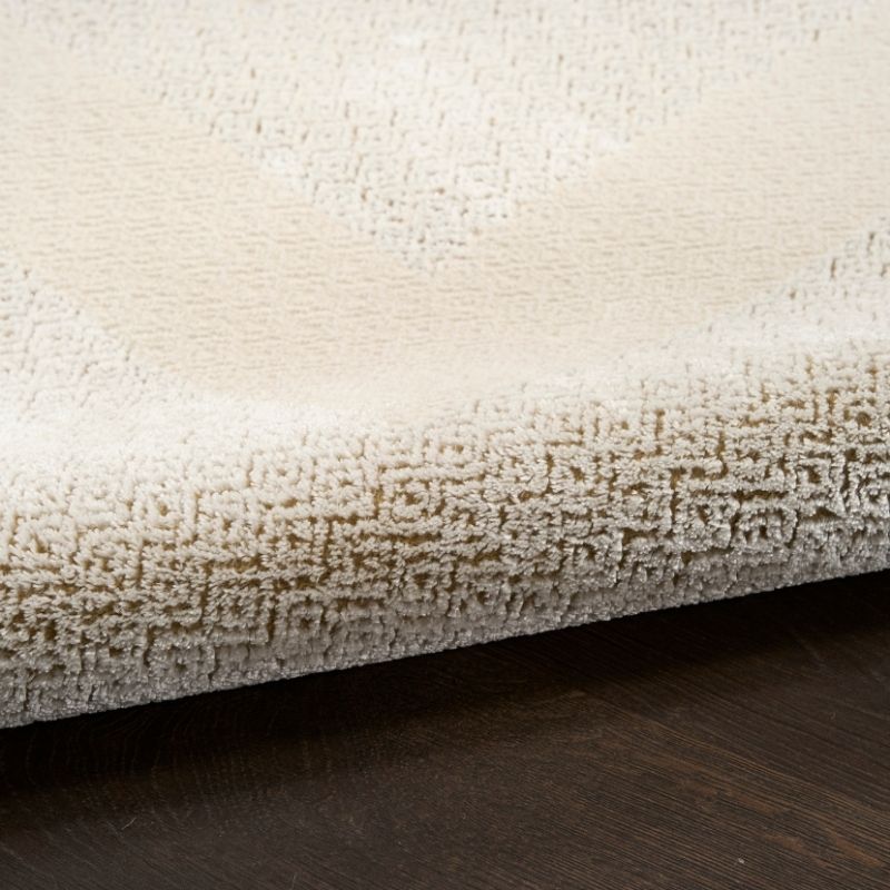 Close-up of a plush cream-colored area rug with a textured loop pile and a subtle geometric pattern, shown folded over to reveal its thickness and underside, resting on a dark wood floor.
