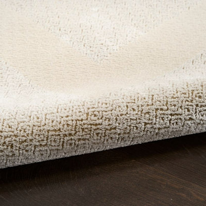 Close-up of a plush cream-colored area rug with a textured loop pile and a subtle geometric pattern, shown folded over to reveal its thickness and underside, resting on a dark wood floor.