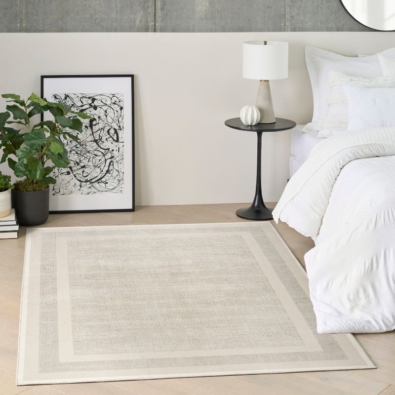 A rectangular area rug in a neutral beige and cream tone with a subtle textured pattern and double border, styled in a modern bedroom next to a bed with white linens, a black side table, and a potted plant.