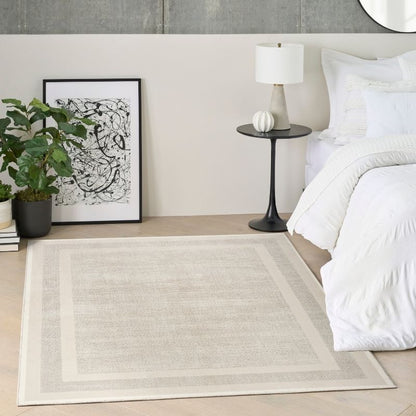 A rectangular area rug in a neutral beige and cream tone with a subtle textured pattern and double border, styled in a modern bedroom next to a bed with white linens, a black side table, and a potted plant.