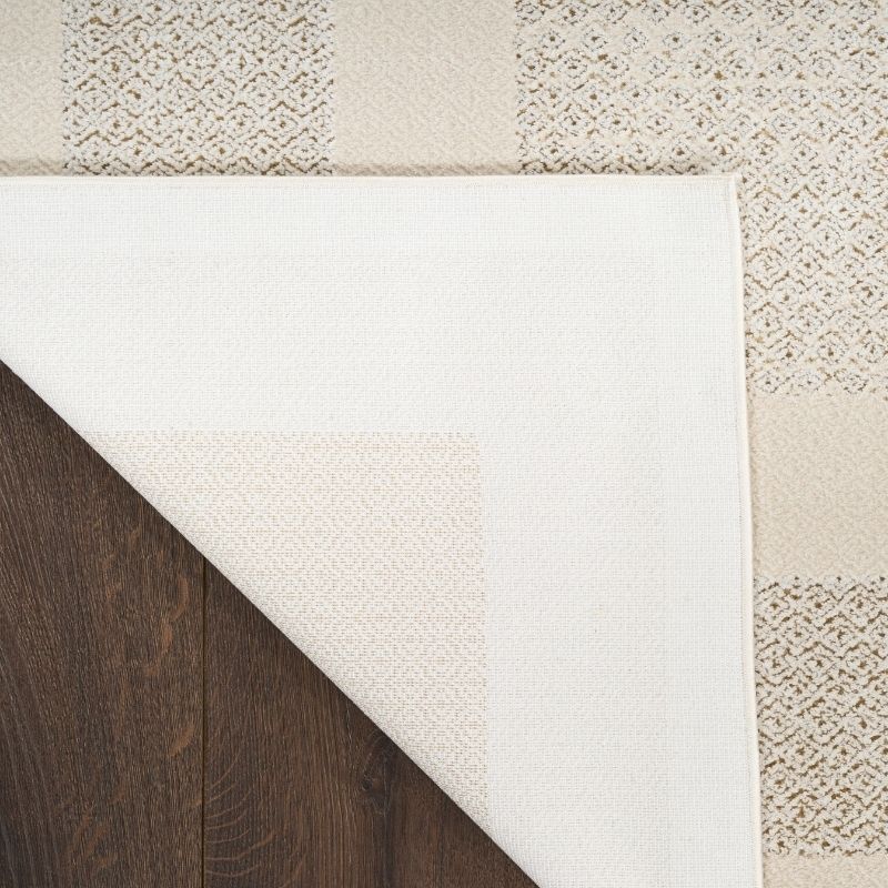 A close-up of a cream and beige patterned rug with one corner folded over to reveal a dark brown wood floor underneath.