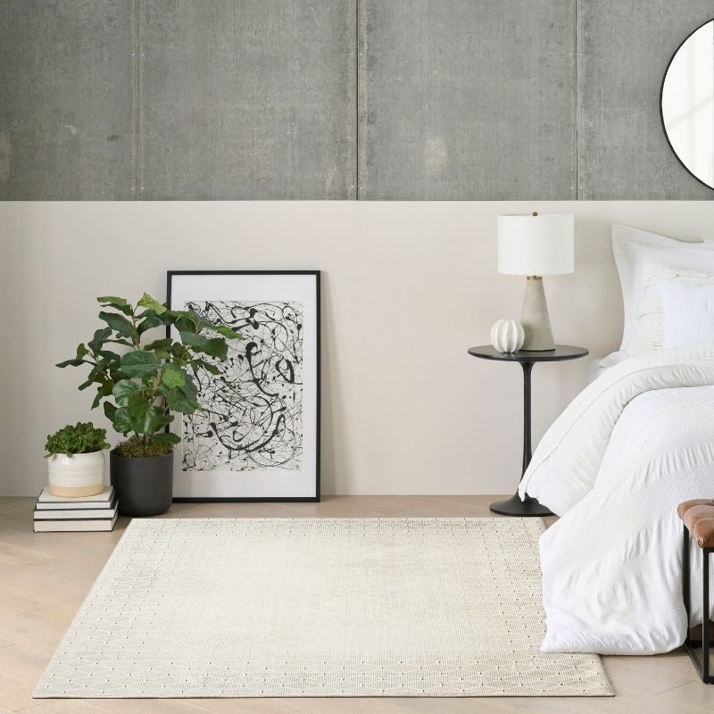A styled bedroom scene featuring a large cream-colored textured area rug on a light wood floor, with a bed, side table, lamp, and potted plant against a two-tone wall.