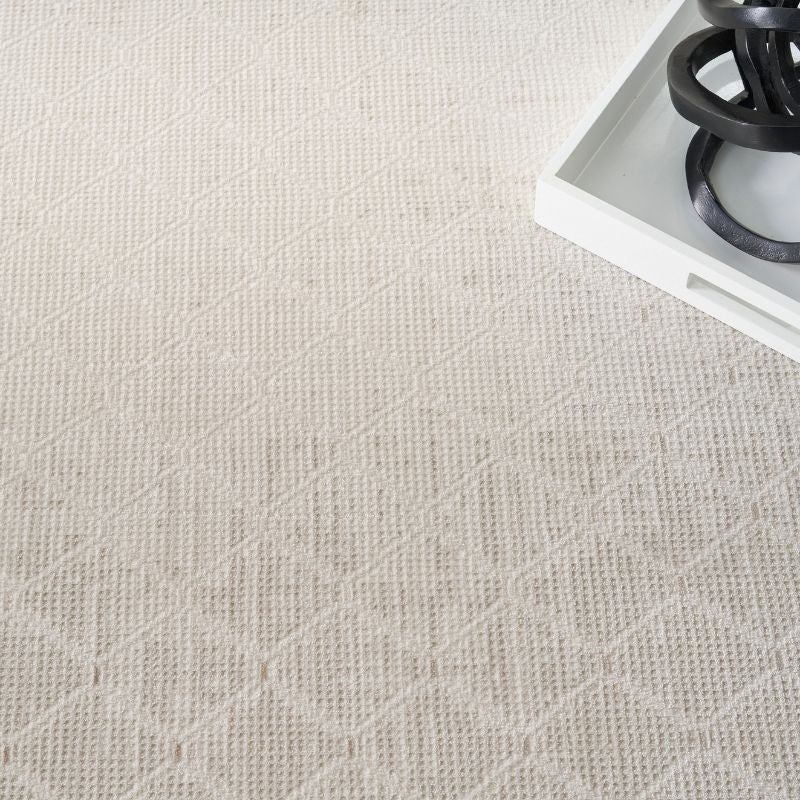 A close-up of a light beige textured rug with a subtle geometric diamond pattern, shown with a white tray and black decorative rings in the top right corner.