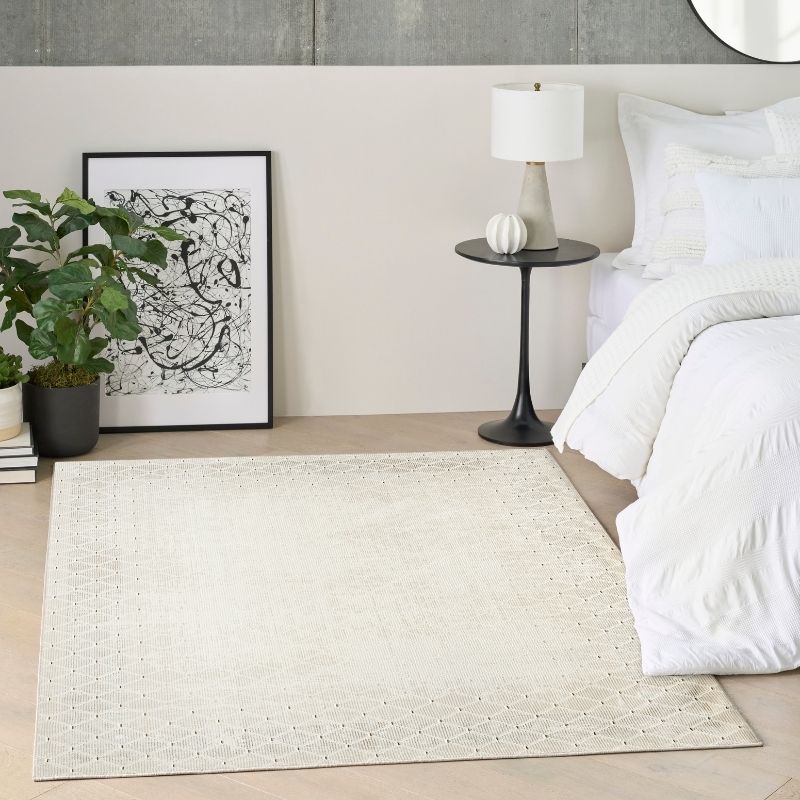 A large rectangular area rug in a warm off-white or cream tone with a subtle woven geometric pattern, styled in a modern bedroom next to a bed with white bedding, a black side table, and a potted plant.
