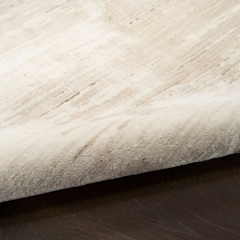 A close-up of a plush, textured area rug in a warm cream or light beige color, shown folded over to reveal its pile and underside, resting on a dark wood floor.