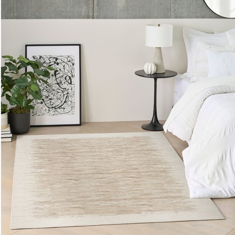 A rectangular area rug in a warm beige and cream textured pattern, styled in a modern bedroom setting next to a bed with white bedding, a black side table with a lamp, and a potted plant.