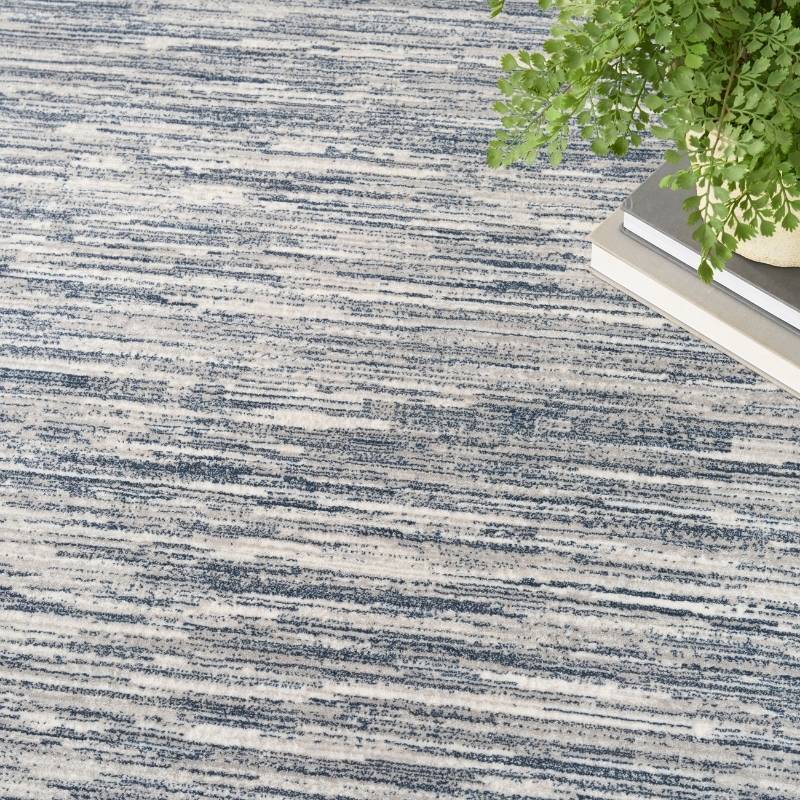 A close-up of a textured area rug with a distressed, horizontal brushstroke pattern in navy blue and off-white. A green potted plant and a stack of books are styled in the top-right corner as props.