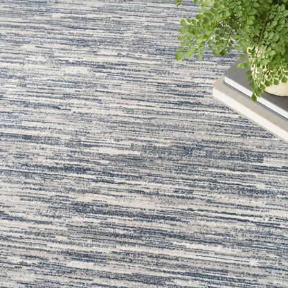 A close-up of a textured area rug with a distressed, horizontal brushstroke pattern in navy blue and off-white. A green potted plant and a stack of books are styled in the top-right corner as props.
