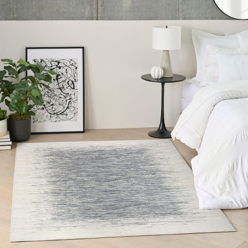 A rectangular area rug with a distressed gray and off-white abstract pattern, styled in a modern bedroom next to a bed and a potted plant.
