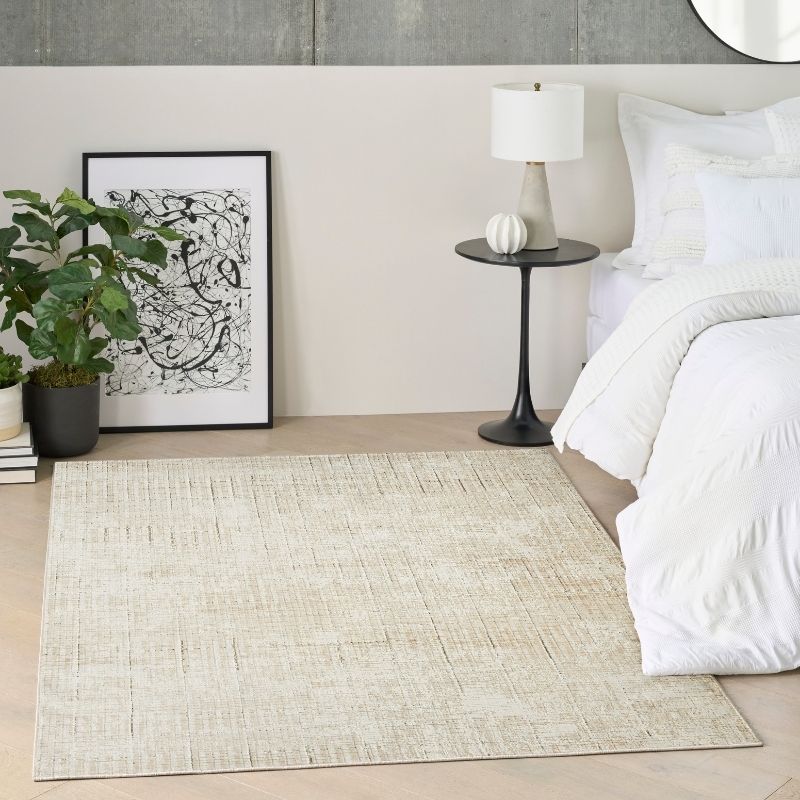 A rectangular area rug in a light beige and off-white textured weave, styled in a modern bedroom setting next to a bed with white linens, a black side table with a lamp, and a potted plant.