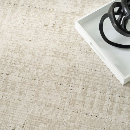 A close-up of a light beige and off-white textured rug with a distressed, woven pattern, styled with a white tray and black decorative rings in the upper right corner.