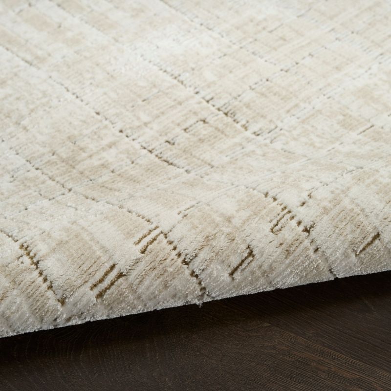 A close-up of a textured area rug in cream and beige with a subtle woven geometric pattern, shown folded over to reveal its underside resting on a dark wood floor.