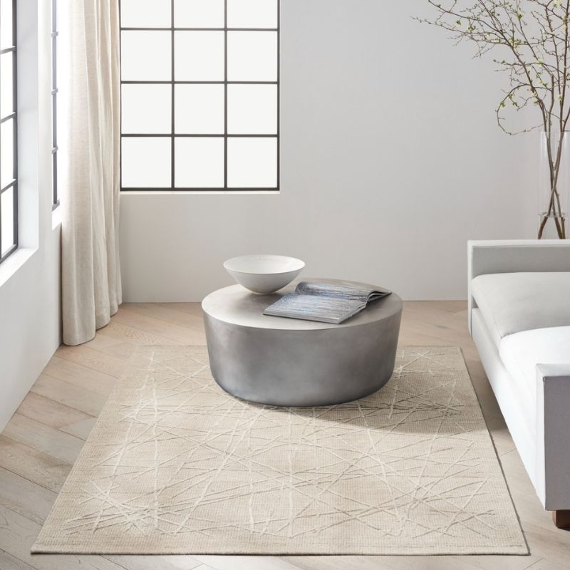 A modern living room scene featuring a large beige area rug with a subtle geometric line pattern. A round, metallic silver coffee table sits on the rug, styled with a white ceramic bowl and a folded gray blanket.