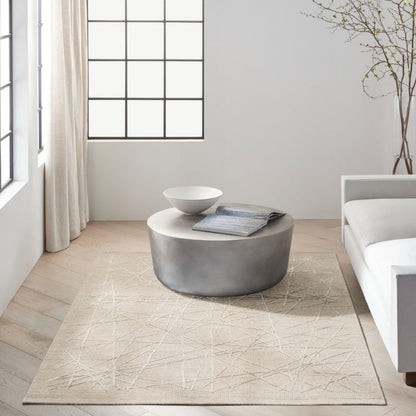 A modern living room scene featuring a large beige area rug with a subtle geometric line pattern. A round, metallic silver coffee table sits on the rug, styled with a white ceramic bowl and a folded gray blanket.