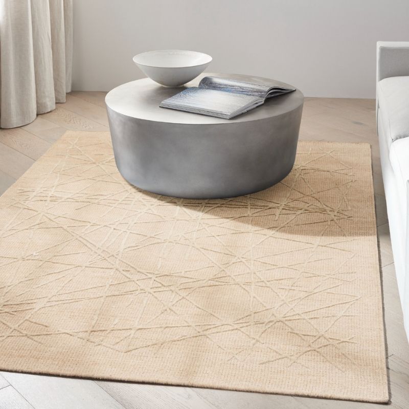 A large beige area rug with a textured geometric line pattern, styled in a modern living room setting under a round gray coffee table.