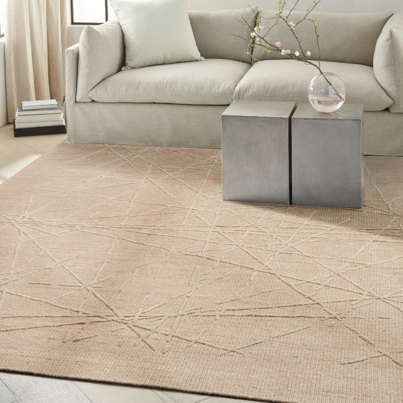 A large beige area rug with a subtle geometric line pattern is styled in a living room setting, positioned under a light gray sofa and a two-tone gray side table.