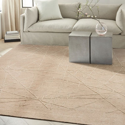 A large beige area rug with a subtle geometric line pattern is styled in a living room setting, positioned under a light gray sofa and a two-tone gray side table.