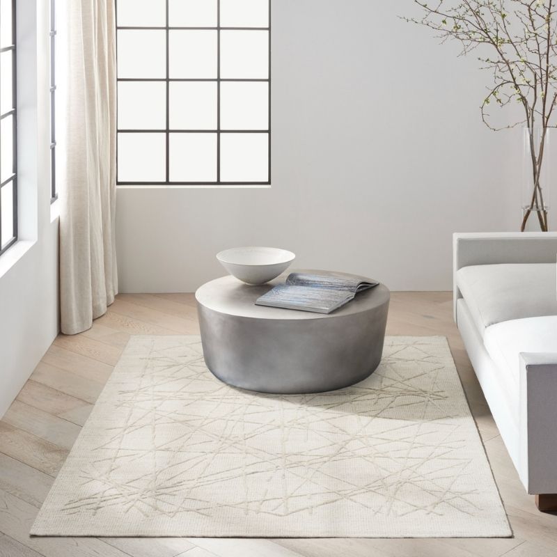 A round gray coffee table in a bright, modern living room setting with a light beige patterned area rug underneath, a white bowl, and a folded magazine on top.