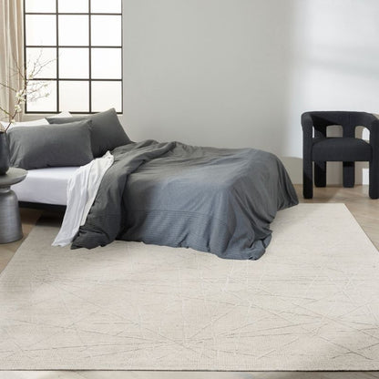 A styled bedroom scene showing a bed dressed in dark gray bedding, placed on a large light beige rug with a subtle geometric line pattern. The room has a white wall, a window with black grid panes, a dark chair, and a small side table.