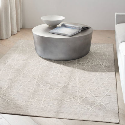 A light beige area rug with a white geometric line pattern, styled in a modern living room setting under a round concrete coffee table.