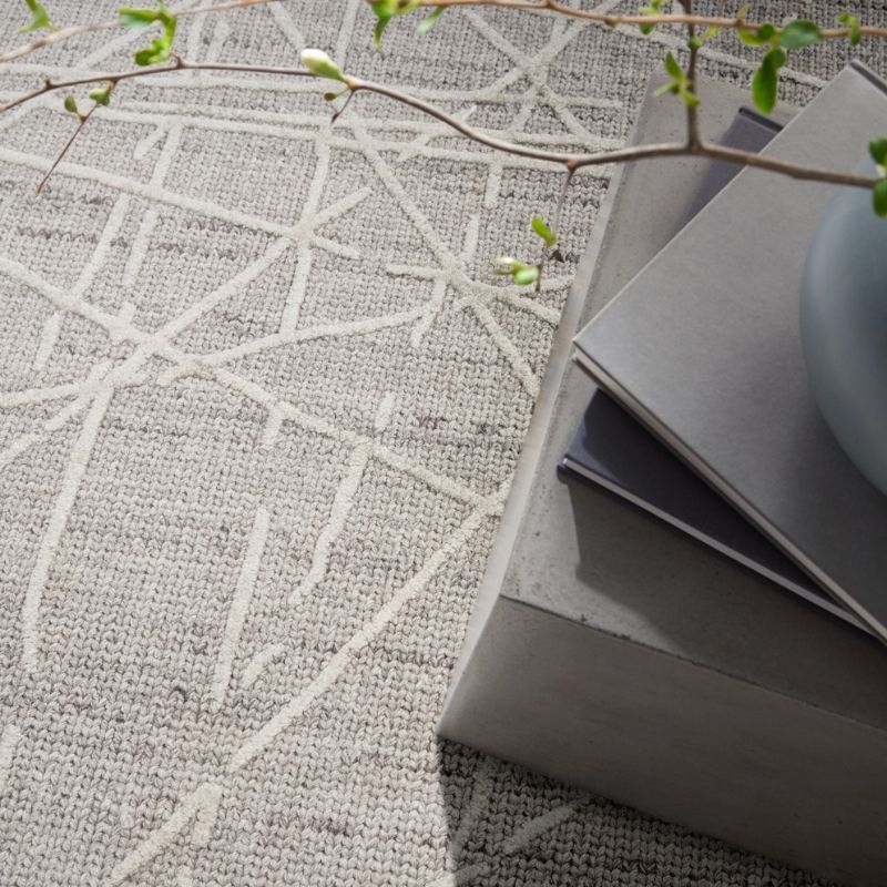 A close-up of a woven rug with a light beige and off-white geometric line pattern, styled with stacked gray books and a pale blue ceramic vase under soft natural light.