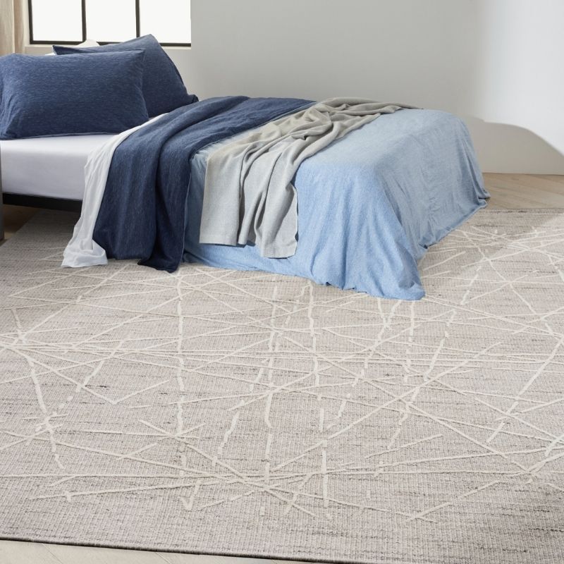 A large light beige area rug with a white geometric line pattern, shown in a bedroom setting under a bed with blue and gray bedding.