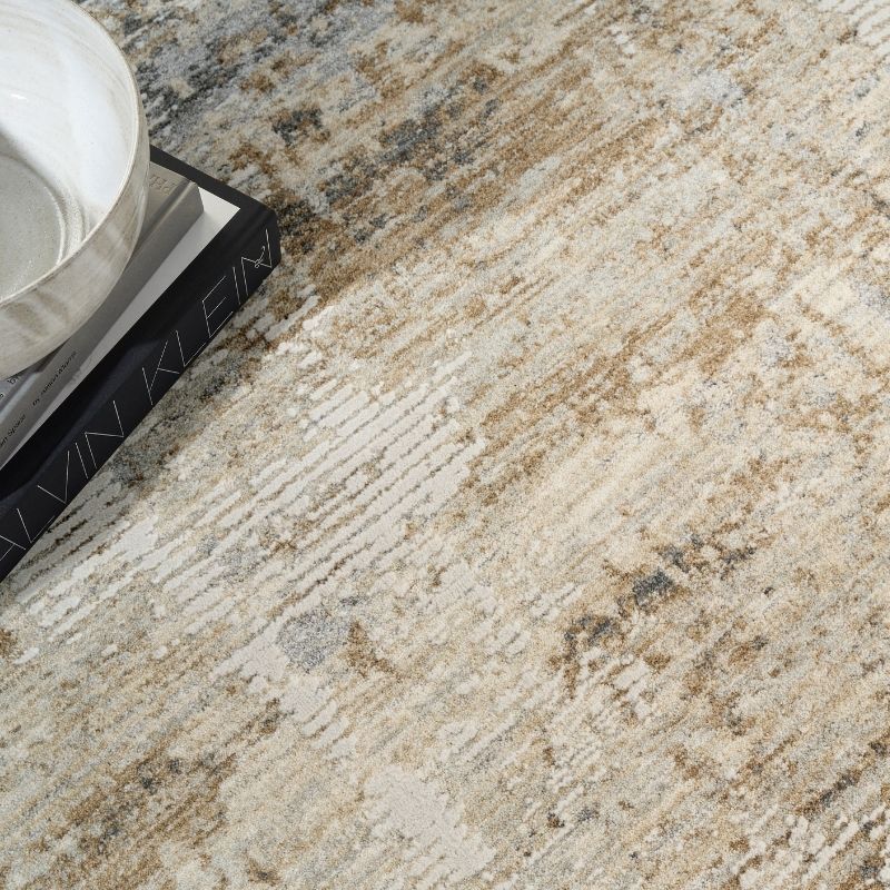 A close-up of a distressed area rug with an abstract pattern in beige, cream, gray, and brown tones, styled with a white ceramic bowl and a black book in the upper left corner.