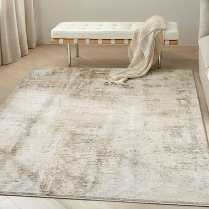A large rectangular area rug with an abstract distressed pattern in cream, beige, and light gray tones, styled in a bright bedroom setting with a bench and throw blanket.