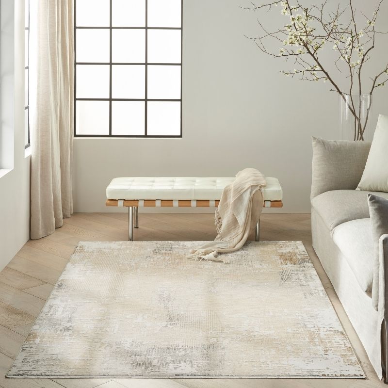 A large rectangular area rug in a neutral cream, beige, and light gray distressed pattern, styled in a bright, modern living room next to a sofa and a bench.