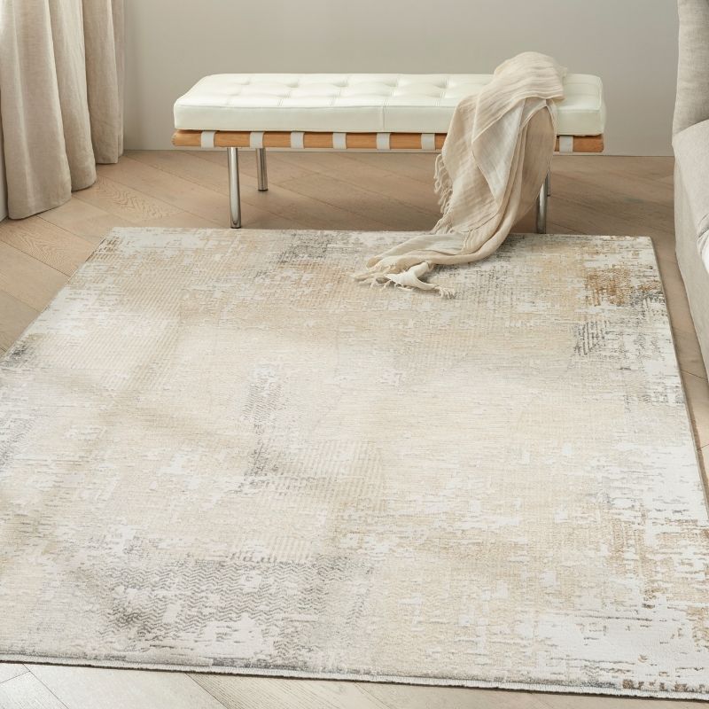 A large rectangular area rug with a distressed, abstract pattern in cream, beige, and light gray tones, styled in a bright room with a tufted bench and draped throw blanket.