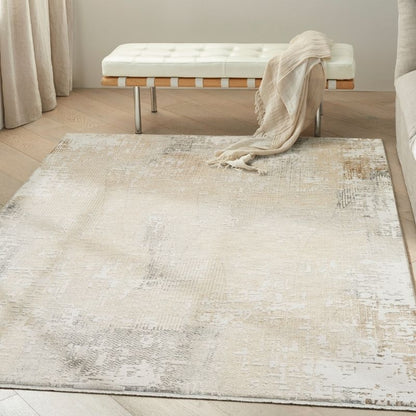 A large rectangular area rug with a distressed, abstract pattern in cream, beige, and light gray tones, styled in a bright room with a tufted bench and draped throw blanket.