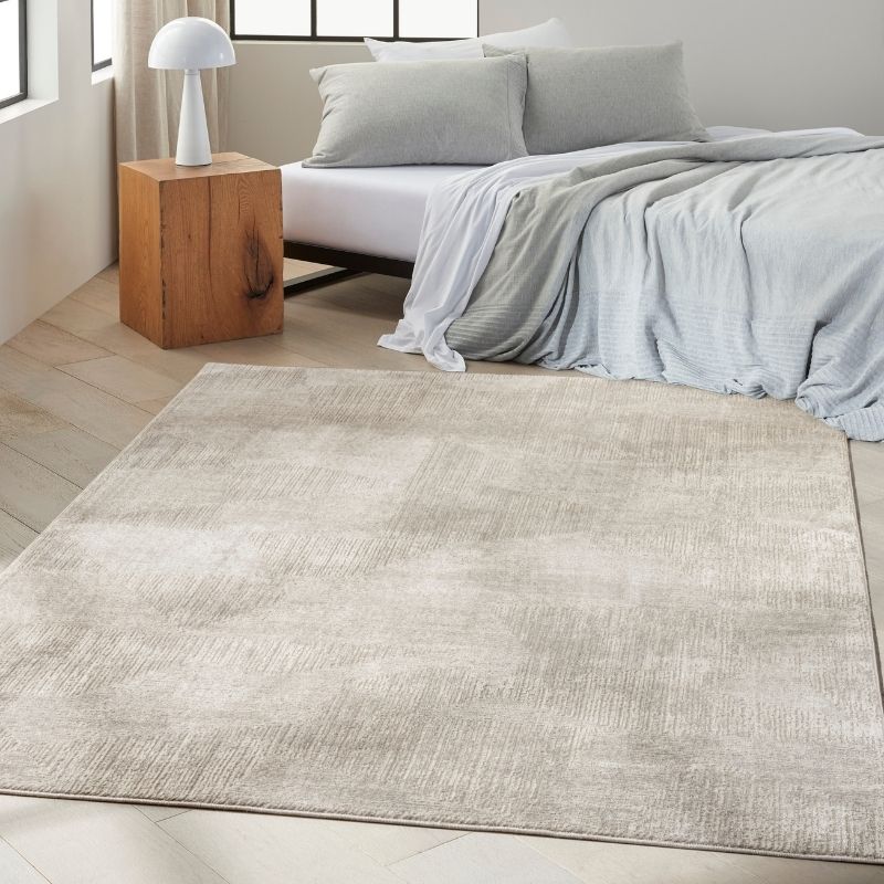 A large rectangular area rug in a light beige and gray textured pattern, shown in a modern bedroom setting next to a bed with gray bedding and a wooden side table.