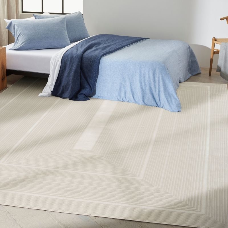 A large cream-coloured area rug with a subtle geometric pattern, shown in a styled bedroom setting under a bed with light blue and navy bedding.