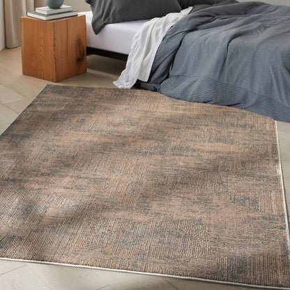 A rectangular area rug with a distressed, textured pattern in shades of brown, beige, and gray, shown on a light-colored floor in a bedroom setting next to a bed.