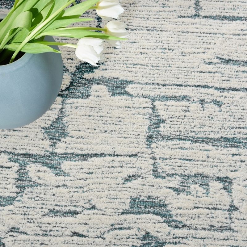 A close-up of a textured area rug with a distressed abstract pattern in muted blue-gray and off-white, styled with a gray ceramic pot and white tulips in the top-left corner.