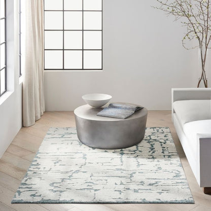 A modern living room scene featuring a rectangular area rug with a distressed abstract pattern in light blue and off-white. A round gray ottoman sits on the rug, styled with a white bowl and a folded blue-gray throw blanket.