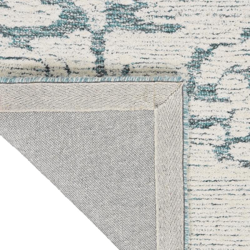 Close-up of a textured area rug corner showing a cream and teal abstract pattern with a light gray underside and fringed edge.