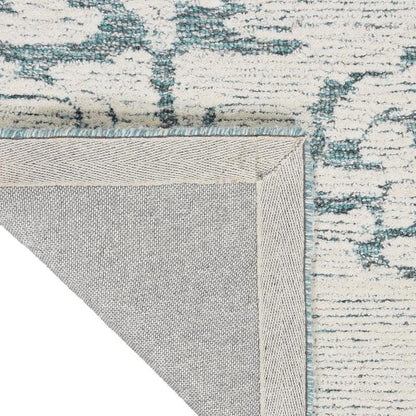 Close-up of a textured area rug corner showing a cream and teal abstract pattern with a light gray underside and fringed edge.