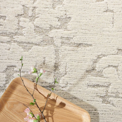 A close-up of a textured area rug with a light beige and gray abstract pattern, styled with a light wood tray and pink cherry blossom branches in the lower left corner.