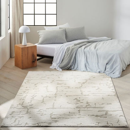A rectangular area rug with a distressed, abstract gray pattern on a cream base, styled in a bright, modern bedroom next to a bed with light gray bedding.