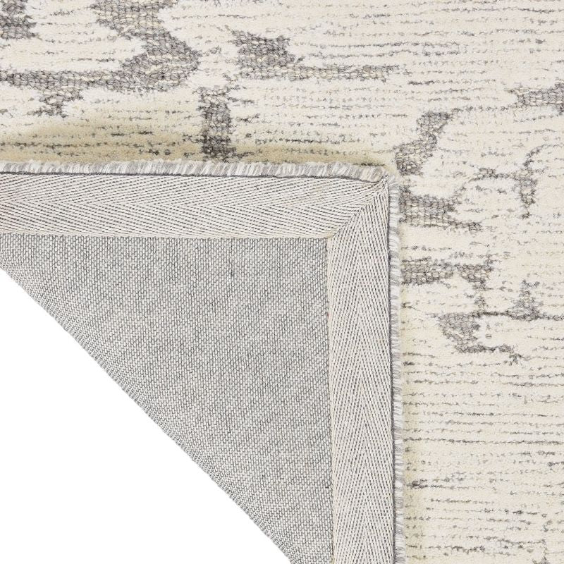 Close-up of a rug corner showing the cream and gray distressed pattern on top and the light gray underside with fringed edge.